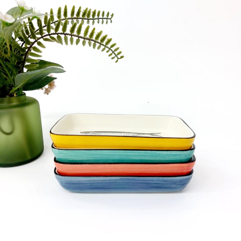 Pode incluir: Uma pilha de quatro travessas retangulares de cer&acirc;mica nas cores amarelo, verde-azulado, coral e azul. Cada travessa tem uma borda preta e um interior branco. A travessa superior apresenta uma ilustra&ccedil;&atilde;o de peixe. Um vaso verde com vegeta&ccedil;&atilde;o est&aacute; no fundo.