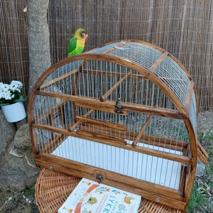 May include: A wooden birdcage with a metal wire mesh. The cage has a rounded top and a hinged door. A green and yellow parakeet is perched on the top of the cage.