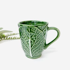Könnte beinhalten: Eine grüne Keramiktasse mit einem Kohlblatt-Design. Die Tasse hat einen Henkel und ist etwa 10 cm hoch. Die Tasse hat eine leuchtend grüne Farbe und eine glänzende Oberfläche.