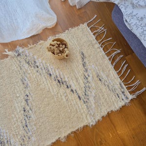 Puede incluir: Una alfombra rectangular beige tejida con una superficie texturizada y un borde con flecos. La alfombra presenta un patrón en zigzag azul oscuro y una pequeña cesta tejida llena de flores secas. La alfombra está sobre un suelo de madera.