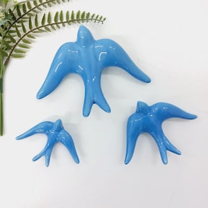 Könnte beinhalten: Drei glänzende, hellblaue Keramikschwalben in verschiedenen Größen. Die Vögel sind mit ausgebreiteten Flügeln dargestellt, was auf Flug hindeutet. Die glatte, reflektierende Oberfläche der Keramik verleiht ihnen einen modernen, dekorativen Reiz. Ein Zweig grünes Laub ist oben links sichtbar.