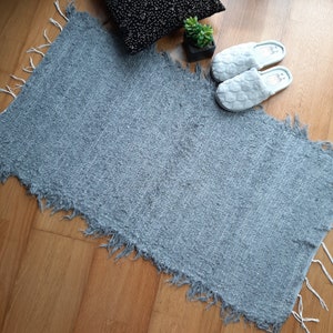May include: A light gray, fluffy rug with a frayed edge. The rug is laid out on a wooden floor.