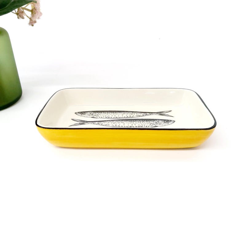 Pode incluir: Uma travessa retangular de cer&acirc;mica com exterior amarelo brilhante e interior branco. Duas ilustra&ccedil;&otilde;es detalhadas de peixes em preto e branco est&atilde;o centradas dentro da travessa. A borda &eacute; delineada com uma fina borda preta.