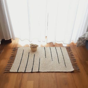 Peut inclure: Un tapis tissé blanc avec des rayures noires et des franges brunes. Le tapis est sur un sol en bois.