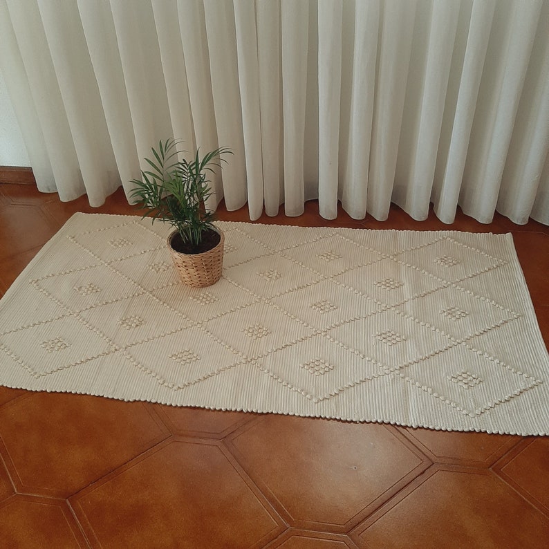 May include: A white woven rug with a diamond pattern. The rug is on a wooden floor and a potted plant is in the foreground.