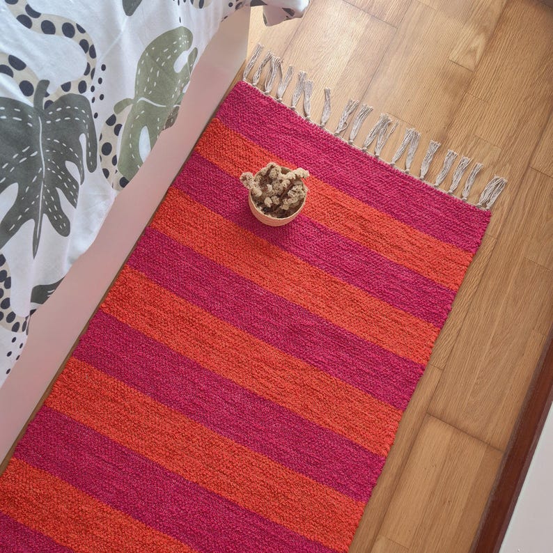 May include: A rectangular rug with alternating horizontal stripes of bright pink and orange. The rug has a fringed edge and is placed on a wooden floor. A small wooden bowl with dried flowers sits on the rug.