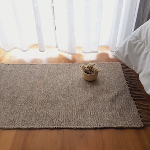 Pode incluir: Um tapete retangular bege tecido com bordas franjadas. Uma pequena cesta tecida cheia de flores secas está sobre o tapete. O tapete é colocado em um piso de madeira, com uma cortina branca transparente ao fundo.