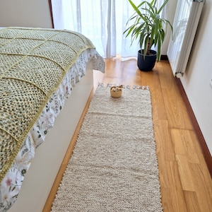 Op de afbeelding: Een rechthoekig, lichtbeige geweven vloerkleed met een getextureerd oppervlak. Het vloerkleed ligt op een houten vloer naast een bed met een groen en wit patroon. Een klein geweven mandje staat op het vloerkleed.