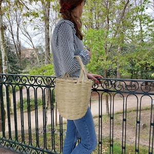 K&ouml;nnte beinhalten: Eine Frau in einem gestreiften Shirt und blauen Jeans tr&auml;gt eine geflochtene Strohtasche.