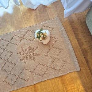 Può includere: Un tappeto rettangolare beige caratterizzato da un motivo a stella centrale e forme a diamante circostanti create con punti trapuntati in rilievo. Un vaso in ceramica bianco a forma di cuore con fiori secchi è posizionato sul tappeto.