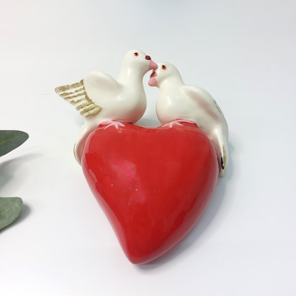 May include: Two white ceramic doves are perched on a red heart-shaped base. The doves are facing each other and appear to be kissing.