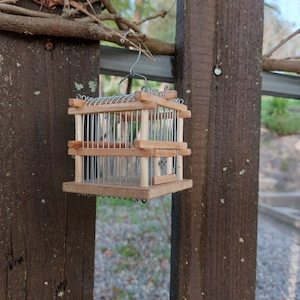 Pu&ograve; includere: Una piccola gabbia per uccelli in legno con una rete metallica all'interno &egrave; appesa a una recinzione in legno. La gabbia &egrave; in legno marrone chiaro e ha un design semplice.