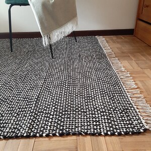 May include: Black and white woven rug with a geometric pattern and fringe edges. The rug is on a wooden floor.