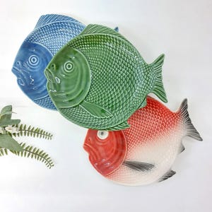 May include: Three ceramic fish-shaped plates in shades of blue, green, and red. The plates have textured scales and detailed fins. The plates are arranged on a white surface, showcasing their vibrant colors and unique design.