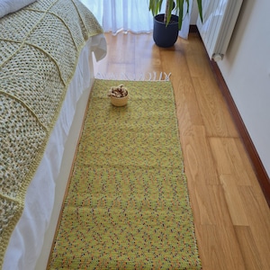 Pode incluir: Um tapete retangular verde limão com um padrão tecido e manchas coloridas. O tapete tem uma borda com franjas e é colocado em um piso de madeira. Uma pequena tigela tecida com itens decorativos está no tapete.