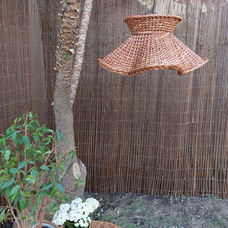May include: A woven wicker lampshade with a flared, bell-like shape. The lampshade is suspended from a black cord and has a natural brown color. The background includes a bamboo fence, a tree trunk, and greenery.
