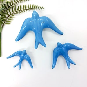 Puede incluir: Tres figuras de cerámica azul claro brillante de golondrinas con las alas extendidas. Las aves varían en tamaño, la más grande en el centro y dos más pequeñas debajo. Las golondrinas están dispuestas sobre una superficie blanca, sugiriendo un tema decorativo.