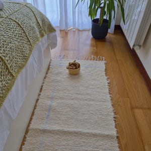 Puede incluir: Una alfombra rectangular de color blanco roto con rayas azul claro y bordes deshilachados. Está colocada sobre un suelo de madera, cerca de una cama con una manta verde. Una pequeña cesta tejida con flores secas se encuentra sobre la alfombra.
