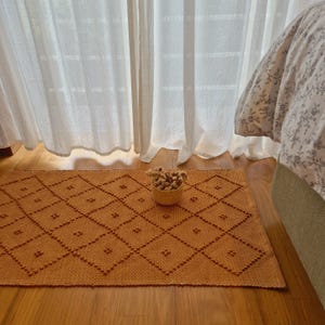 May include: An orange rectangular rug with a diamond pattern and small, raised dots. A small woven basket filled with dried flowers sits on the rug. The rug is placed on a wooden floor near a bed and sheer curtains.
