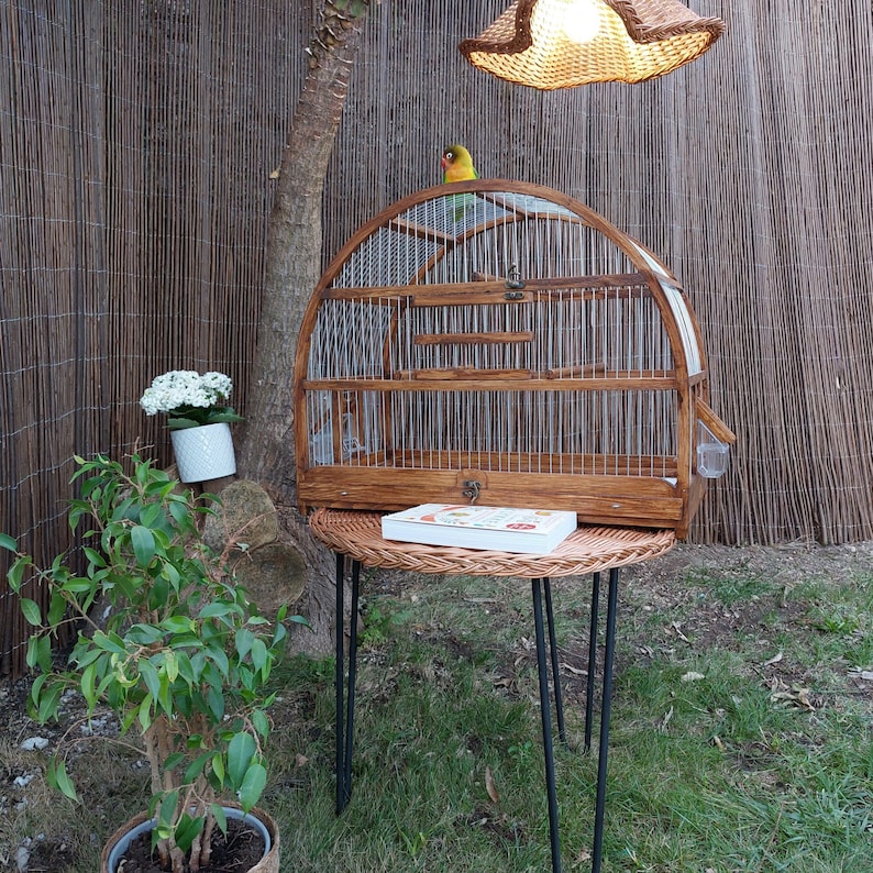 May include: A wooden birdcage with a domed top sits on a wicker table with black metal legs. A small bird is perched on the top of the cage. The cage has a small wooden drawer on the bottom.