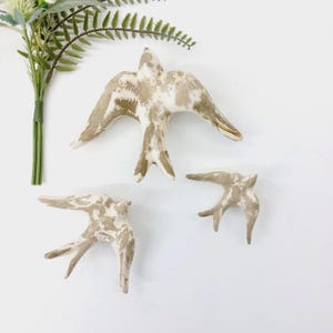 Può includere: Tre decorazioni da parete in ceramica a forma di uccelli in volo, caratterizzate da una finitura screziata bianca e beige dall'aspetto vissuto. Gli uccelli sono disposti in gruppo con le ali spiegate, mostrando una superficie ruvida.