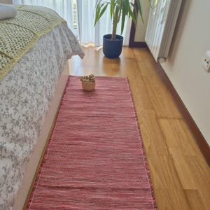 Può includere: Un tappeto passatoia rettangolare caratterizzato da una trama tessuta con un mix di fili rosso scuro e bianchi. Il tappeto presenta una struttura a tessitura piatta con linee orizzontali visibili e frange corte sfilacciate sui lati corti.