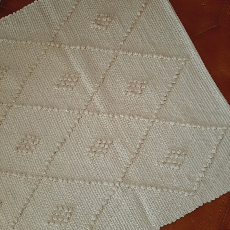 May include: A white woven rug with a diamond pattern. The rug has a textured surface with raised bumps.