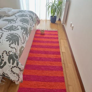 May include: A vibrant, rectangular rug with alternating horizontal stripes of bright pink and orange. The rug is made of a woven material and has a fringed edge. It is placed on a wooden floor next to a bed with a patterned comforter.