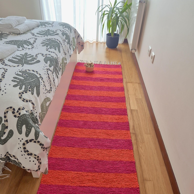 May include: A vibrant, rectangular rug with alternating horizontal stripes of bright pink and orange. The rug is made of a woven material and has a fringed edge. It is placed on a wooden floor next to a bed with a patterned comforter.