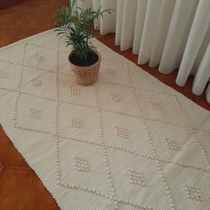 May include: A white woven rug with a diamond pattern. The rug has a textured surface and is made of natural fibers. A potted plant sits on the rug.