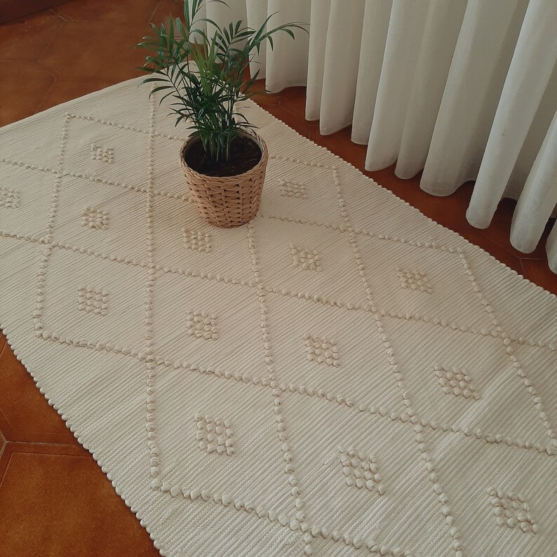 May include: A white woven rug with a diamond pattern. The rug has a textured surface and is made of natural fibers. A potted plant sits on the rug.