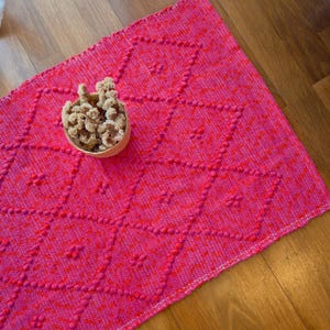 Könnte beinhalten: Ein leuchtend pinkfarbener gewebter Teppich mit einem Rautenmuster und erhabenen, strukturierten Punkten. Ein kleiner geflochtener Korb mit getrockneten Blumen liegt auf dem Teppich. Die Farbe des Teppichs ist ein helles, auffälliges Pink.