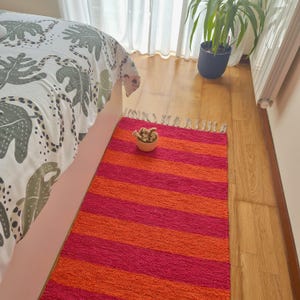 May include: A vibrant, striped rug in shades of red and orange. The rug is rectangular with horizontal stripes and a fringed edge. A small, woven bowl with a decorative item sits on the rug near a bed with a patterned comforter.