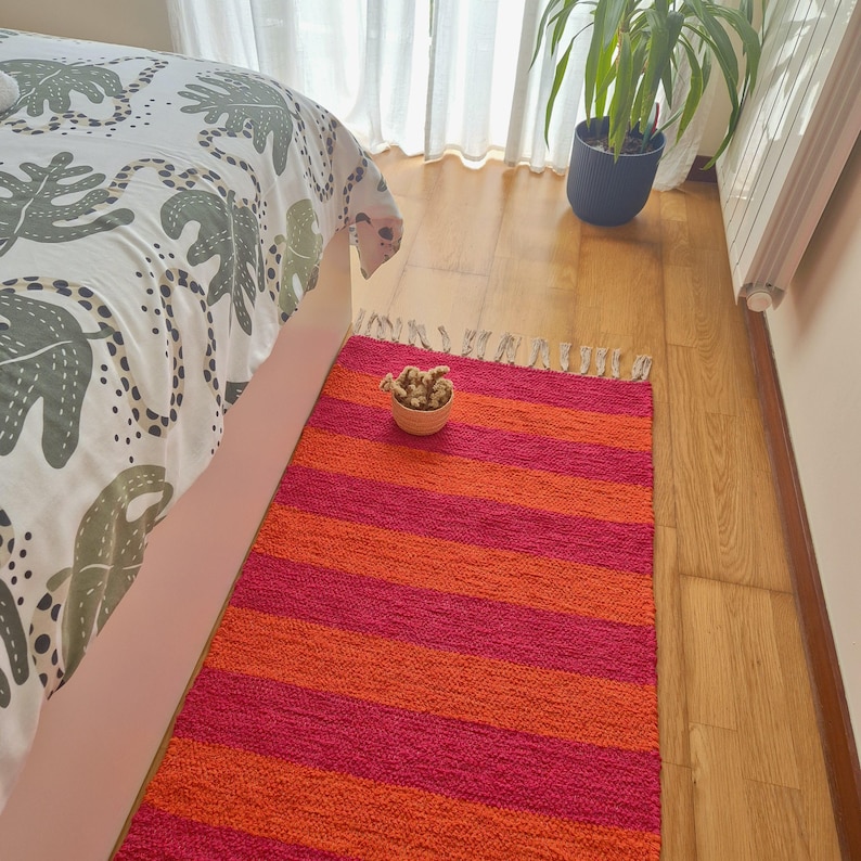 May include: A vibrant, striped rug in shades of red and orange. The rug is rectangular with horizontal stripes and a fringed edge. A small, woven bowl with a decorative item sits on the rug near a bed with a patterned comforter.