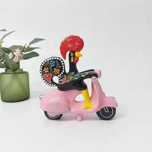 May include: A pink scooter with a black rooster painted on it. The rooster has a red comb and wattle, and its tail feathers are painted with colorful flowers.