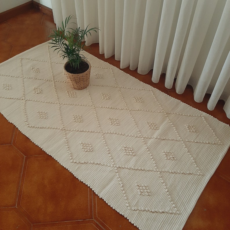May include: A white woven rug with a diamond pattern. The rug has a subtle texture and is made of natural fibers.