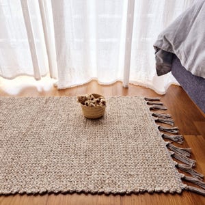 Può includere: Un tappeto rettangolare beige intrecciato con frange su un lato. Un piccolo cestino intrecciato pieno di fiori secchi è appoggiato sul tappeto. Il tappeto è posizionato su un pavimento in legno vicino a un letto e tende trasparenti.