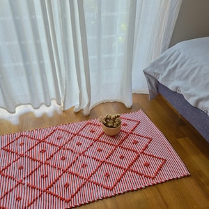 Puede incluir: Una alfombra a rayas rojas y blancas con un diseño de diamantes y detalles de mechones rojos. Un pequeño cuenco tejido lleno de flores secas está colocado sobre la alfombra. La alfombra está sobre un suelo de madera cerca de una ventana con cortinas blancas.