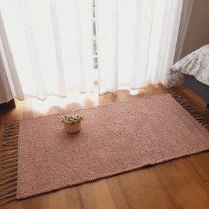 Puede incluir: Una alfombra rectangular de color rosa polvoriento con un tejido texturizado y bordes con flecos. La alfombra está colocada sobre un suelo de madera, con una pequeña cesta tejida de flores secas encima. Cortinas blancas transparentes y una cama en el fondo.