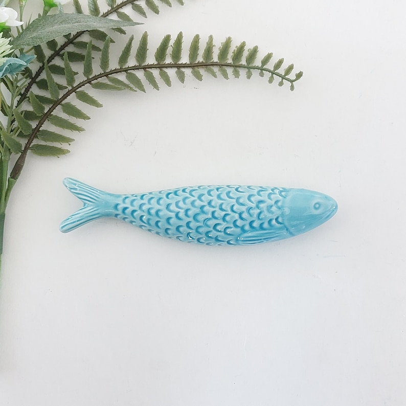 Puede incluir: Una figurita de pescado de cer&aacute;mica azul claro con escamas. El pez mide aproximadamente 13 cm de largo y tiene un cuerpo ligeramente curvado.