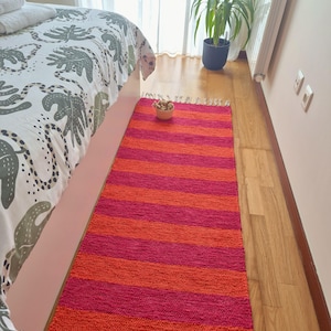 May include: A rectangular, striped rug in shades of orange and fuchsia. The rug is placed on a wooden floor next to a bed with a patterned comforter. A small bowl of dried flowers sits on the rug.