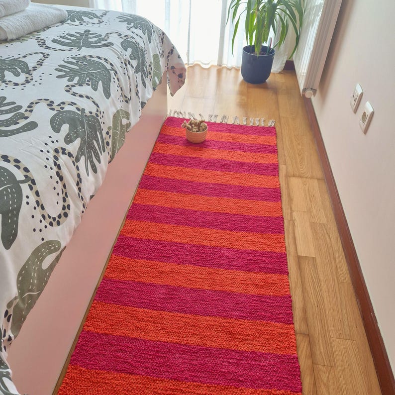 May include: A rectangular, striped rug in shades of orange and fuchsia. The rug is placed on a wooden floor next to a bed with a patterned comforter. A small bowl of dried flowers sits on the rug.