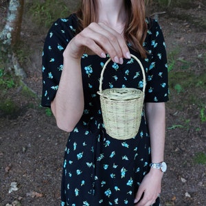 Op de afbeelding: Een vrouw in een zwarte jurk met een bloemenpatroon houdt een klein, geweven mandje met een handvat vast. Het mandje is lichtbruin en rond van vorm.