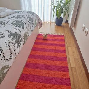 Puede incluir: Una alfombra rectangular con rayas horizontales en tonos naranja y fucsia. La alfombra está colocada sobre un suelo de madera junto a una cama con una colcha estampada. Un pequeño cuenco con objetos decorativos está sobre la alfombra.