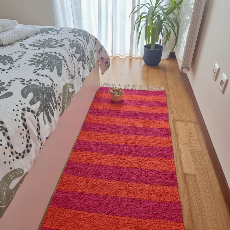 May include: A rectangular rug with horizontal stripes in shades of orange and fuchsia. The rug is placed on a wooden floor next to a bed with a patterned comforter. A small bowl of decorative items sits on the rug.