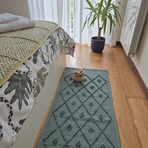 May include: A teal rectangular rug with a diamond pattern and textured details lies on a wooden floor. A small woven basket with beige items sits on the rug. A bed with a patterned comforter and a potted plant are in the background.