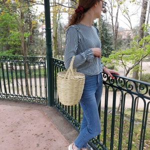 K&ouml;nnte beinhalten: Eine Frau in einem gestreiften blau-wei&szlig;en Oberteil und blauen Jeans h&auml;lt eine geflochtene Strohk&ouml;rbchen-Tasche mit Henkel. Sie steht vor einem schwarzen Metallgel&auml;nder.
