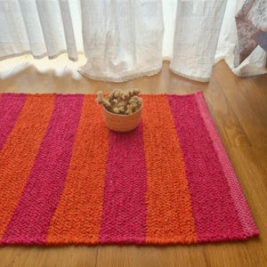 May include: A rectangular rug with vertical stripes in shades of pink and orange. A small woven basket filled with dried flowers sits on the rug. The rug is made of a textured material and is placed on a wooden floor.