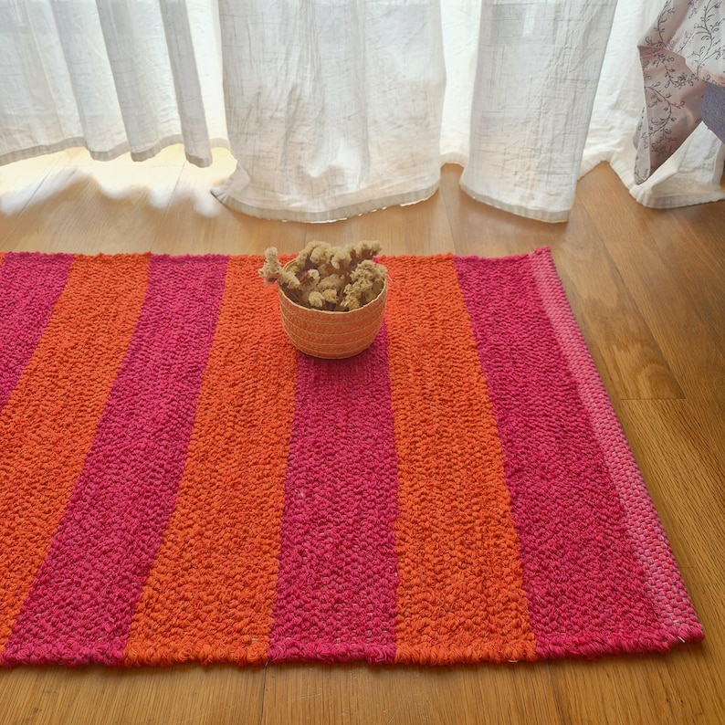 May include: A rectangular rug with vertical stripes in shades of pink and orange. A small woven basket filled with dried flowers sits on the rug. The rug is made of a textured material and is placed on a wooden floor.