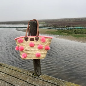 May include: A pink and brown woven tote bag with pink pom poms hanging from the sides. The bag is hanging from a wooden post over a body of water.